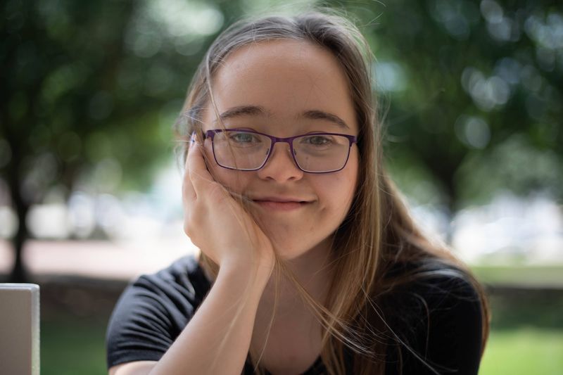 A smiling young woman rests her chin in her hand in a park.
