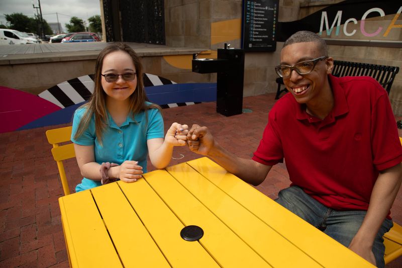 Two friends bump fists and laugh at a yellow cafe table.