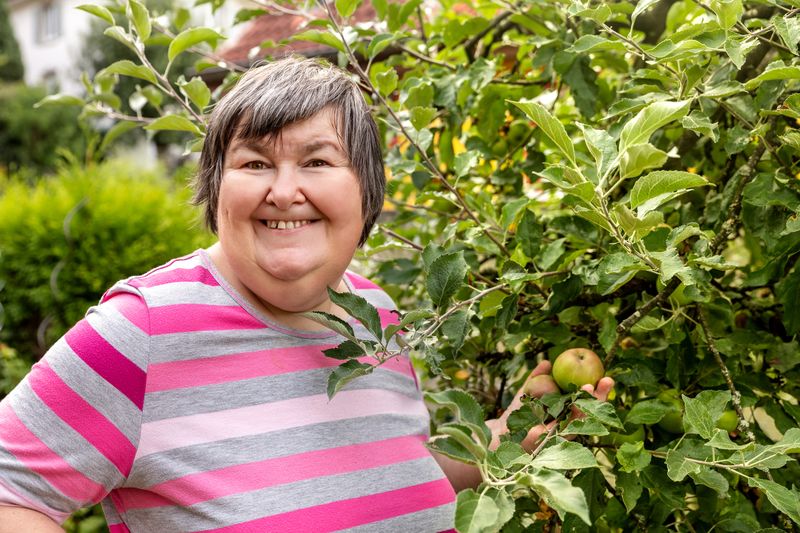 An older woman smiles beside an apple tree in a garden.