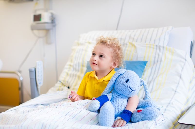 A young child sits up in a hospital bed holding a stuffed animal.