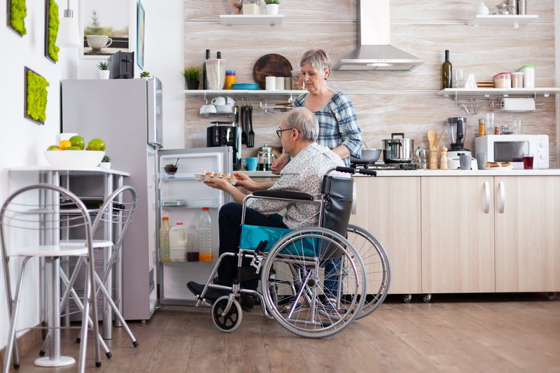 An older man in a wheelchair makes coffee in his kitchen while a woman cooks nearby.