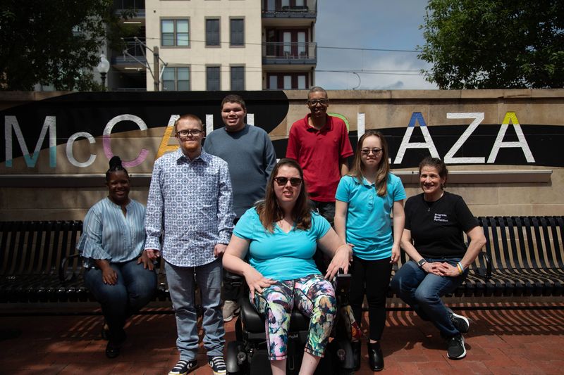 A group of friends, one using a power wheelchair, gathers in a downtown plaza.