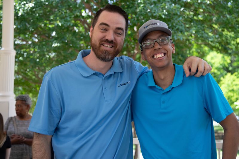 Two friends in blue shirts stand arm in arm and smile.
